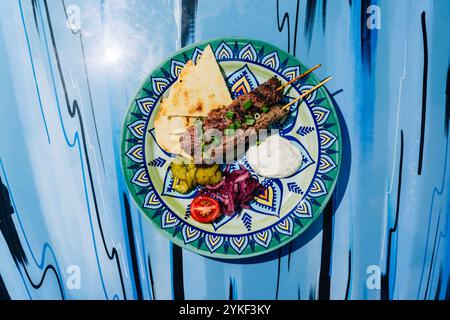 Herzhaftes Rindfleischkebab serviert mit aufgeschnittenem Pita, verschiedenen Gurken, frischen Tomaten und einer cremigen Sauce, wunderschön präsentiert auf einer lebendig gemusterten Platte Stockfoto