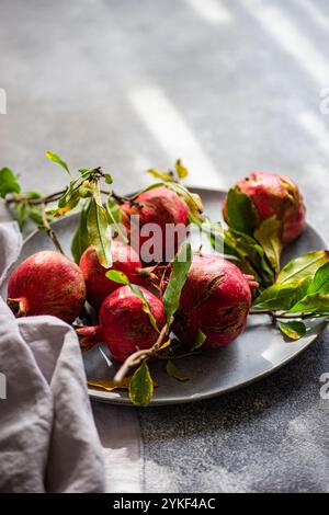 Reife Granatäpfel mit grünen Blättern, die ordentlich auf einer runden grauen Platte angeordnet sind und durch natürliches Licht auf einer strukturierten Oberfläche hervorgehoben werden. Stockfoto