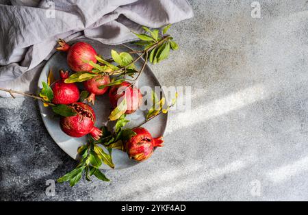 Blick von oben auf eine Gruppe reifer Granatäpfel, einige in Scheiben geschnitten, angeordnet auf einem grauen Steingut-Teller neben einem drapierten grauen Tuch, gegen ein strukturiertes g gestellt Stockfoto