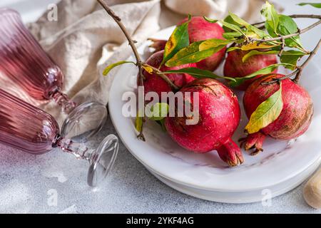 Dunkelrote reife Granatäpfel mit leuchtend grünen Blättern, angeordnet auf einer weißen dekorativen Platte neben eleganten rosafarbenen Glaswaren, auf einem strukturierten Hintergrund Stockfoto