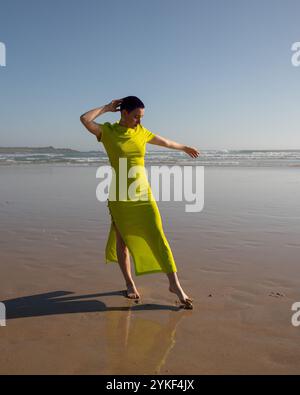 Eine junge Frau mit kurzen violetten Haaren in einem leuchtend gelben Kleid macht einen einsamen Spaziergang entlang der Sandküste eines Strandes. Sie hat die Augen zu Stockfoto