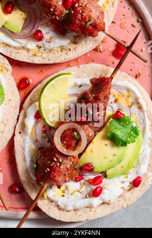 Blick von oben auf köstliche Spieße, serviert mit Joghurtsauce auf Naan-Brot, garniert mit geschnittener Avocado, Granatapfelkernen, roten Zwiebelringen und Sesamsamen Stockfoto