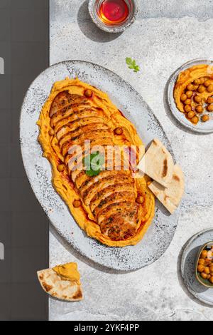 Blick von oben auf ein Gourmet-Menü mit dünn geschnittenem, geröstetem Huhn, elegant auf cremigem Hummus, garniert mit Kräutern und Kichererbsen, begleitet Stockfoto