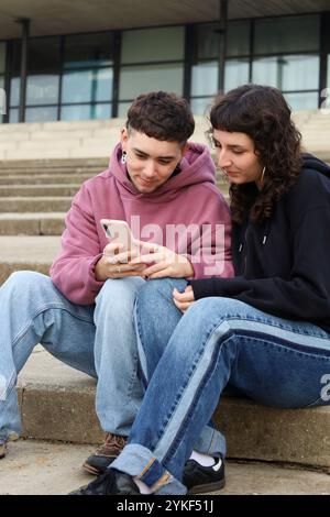 Charmantes LGBTQIA+-Paar sitzt dicht auf Betonstufen im Freien, während sie Inhalte auf einem Smartphone teilen, einen Transmann hervorheben und PR feiern Stockfoto
