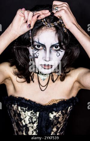 Eine Frau hat eine gotische Ästhetik und zeigt dramatisches Make-up mit schwarz-weißer Gesichtsfarbe und dunklem Lippenstift. Sie hält einen Rosenkranz und trägt dunkles lac Stockfoto