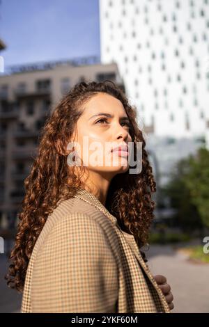 Eine junge brasilianische Geschäftsfrau sieht entschlossen aus, wenn sie draußen in einer Stadt steht, mit Bürogebäuden im Hintergrund und Sonnenlicht, das h hervorhebt Stockfoto