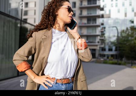 Eine junge brasilianische Geschäftsfrau in einem stylischen Blazer spricht auf ihrem Handy, während sie auf einem Bürgersteig der Stadt steht, ihre lockigen Haare und ihr modernes Outfit reflektieren Stockfoto