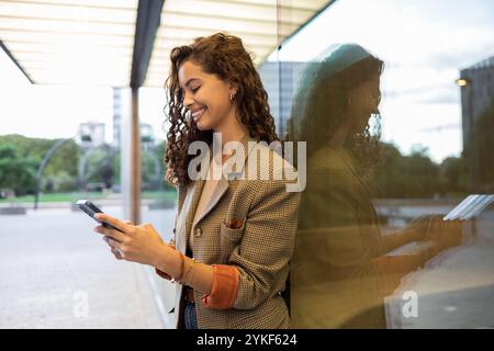 Eine lächelnde brasilianische Geschäftsfrau in professioneller Kleidung verwendet ihr Smartphone, steht an einer Glasfassade in einer städtischen Umgebung und spiegelt Selbstvertrauen wider Stockfoto