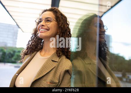 Eine lächelnde brasilianische Geschäftsfrau in einem stilvollen Blazer steht in einem modernen Büro an einem Fenster, ihre Reflexion ist sichtbar und unterstreicht ihr Selbstvertrauen und ihre Selbstvertrauen Stockfoto