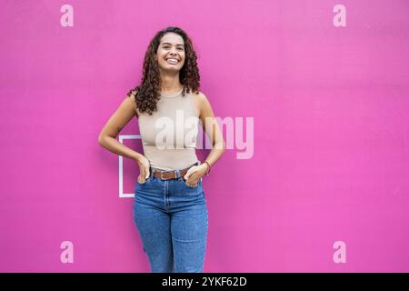 Eine fröhliche junge brasilianische Frau steht selbstbewusst mit der Hand auf der Hüfte vor einem leuchtend rosa Hintergrund Stockfoto