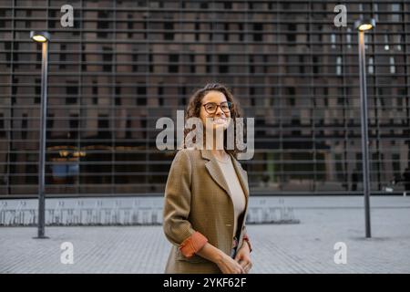 Eine junge brasilianische Geschäftsfrau mit lockigem Haar und Brille, die einen stylischen karierten Blazer trägt, lächelt in einer urbanen Umgebung mit modernem AR Stockfoto