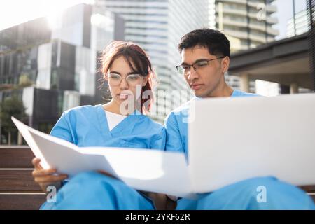 Zwei Schwesternstudenten in blauem Scrubs konzentrieren sich auf das Studium im Freien und konsultieren sowohl einen Laptop als auch Papierdokumente, vor einem modernen urbanen Hintergrund Stockfoto