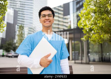 Ein junger Mann in blauer Krankenschwester-Uniform steht draußen mit einem Laptop in der Hand und lächelt selbstbewusst in einer urbanen Umgebung Stockfoto