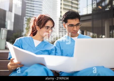 Zwei junge Pflegestudenten, ein Mann und eine Frau, arbeiten mit einem Laptop außerhalb eines modernen Gebäudes an ihren Lehrveranstaltungen zusammen Stockfoto