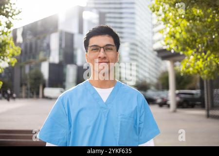Ein junger Schwesternschüler in blauem Gewand lächelt selbstbewusst, steht draußen, mit einem verschwommenen Stadtbild Hintergrund Stockfoto