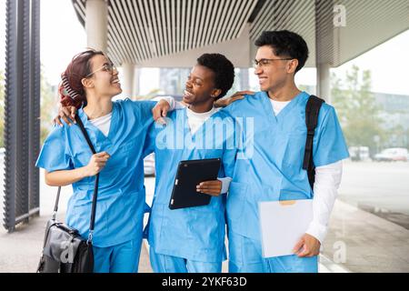 Drei multikulturelle Pflegestudenten in blauen Peelings interagieren gerne und teilen ihre Notizen außerhalb einer medizinischen Einrichtung Stockfoto