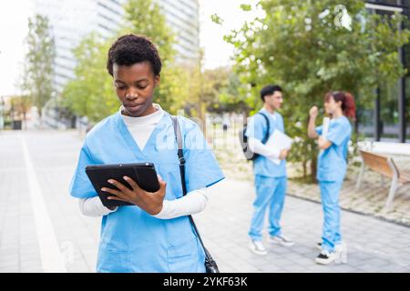 Eine fokussierte junge Frau in einer Krankenschwester-Uniform benutzt ein digitales Tablet, während sie auf einem Universitätsgelände läuft und zwei Kollegen im Hintergrund plaudern Stockfoto