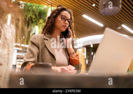 Eine fokussierte brasilianische Geschäftsfrau in Brillen und stilvoller Kleidung Multitasking, während sie auf ihrem Mobiltelefon unterhält und auf einem Laptop arbeitet, in einem modernen, Stockfoto