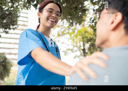 Eine junge Krankenpflegeschülerin in blauem Kleid und Brille lächelt, als sie einem Patienten im Freien hilft und ihm hilft, umgeben von Grün Stockfoto