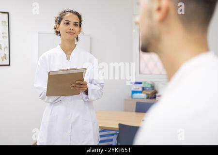 Ein medizinisches Fachpersonal in einem weißen Mantel hält ein Klemmbrett und berät einen Patienten in einer klinischen Umgebung, die die Umgebung reflektiert Stockfoto