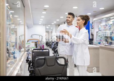 Zwei medizinische Fachkräfte stehen in einem orthopädischen Versorgungsgeschäft, diskutieren Produkte und prüfen Details auf einem Tablet, umgeben von medizinischen Geräten Stockfoto