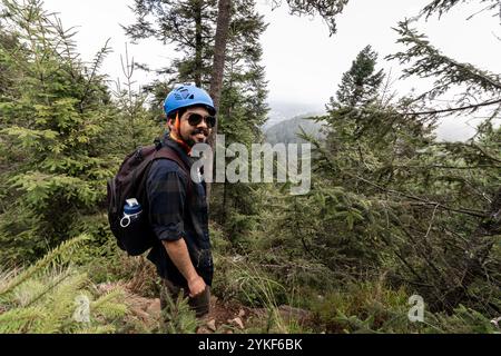 Ein lächelnder Wanderer mit blauem Helm und Sonnenbrille steht zwischen immergrünen Bäumen und trägt einen Rucksack mit einer Wasserflasche Stockfoto