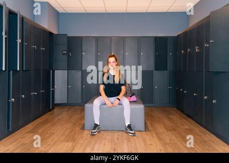 Eine Frau sitzt ruhig in einem Umkleideraum, auf einer Bank zwischen Reihen dunkelblauer Schließfächer. Sie ist in Sportkleidung gekleidet und blickt direkt auf die CA Stockfoto