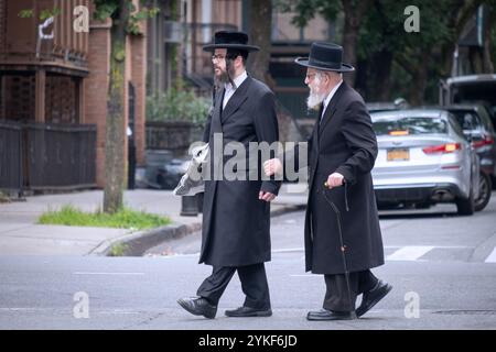 Zwei orthodoxe jüdische Männer, wahrscheinlich Großvater und Enkel, gehen über eine Straße in Brooklyn, zusammen mit dem jüngeren Mann, der Unterstützung leistet. Stockfoto