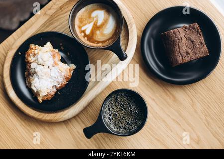 Von oben Frühstücksessen, darunter ein Croissant mit Mandeln, ein dichter Schokoladenbrownie und zwei Kaffeesorten, die wunderschön auf einem präsentiert werden Stockfoto