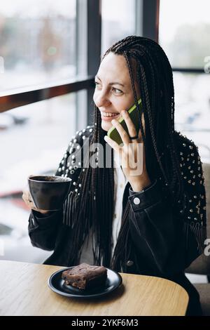 Eine stilvolle kaukasische Frau mit geflochtenen Haaren genießt eine Tasse Kaffee an einem Café-Tisch und lächelt, während sie auf ihrem Handy spricht. Ein Stück Kuchenbegleitung Stockfoto