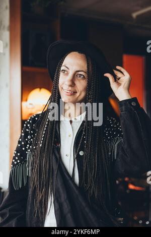 Eine stilvolle weiße Frau mit freundlichem Ausdruck trägt einen schwarzen Hut und Zöpfe, während sie ihren Hut in einem Café verstellt und einen modernen Stadtstil präsentiert Stockfoto