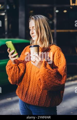 Eine Frau in einem leuchtend orangefarbenen Strickpullover hält eine Kaffeetasse und ein Smartphone und genießt einen sonnigen Tag im Freien mit dem gemütlichen Pullover und dem lässigen Vibe Capture Stockfoto