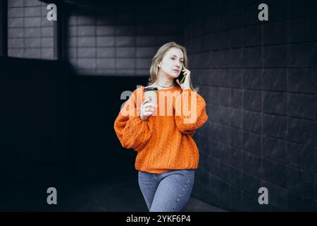 Eine Frau in einem warmen, orangefarbenen Strickpullover genießt einen Kaffee, während sie am Telefon telefoniert. Die Umgebung ist gemütlich und entspannt und sorgt für eine herbstliche Atmosphäre mit ca. Stockfoto