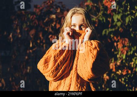 Eine Frau, die einen kuscheligen, orangefarbenen Strickpullover trägt, der teilweise ihr Gesicht bedeckt, steht mitten im leuchtenden Herbstlaub Stockfoto