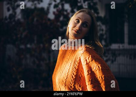 Eine Frau in einem kuscheligen orangefarbenen Pullover steht draußen und lässt sich im warmen Herbstlicht mit ihrem entspannten Auftreten und dem strukturierten Strickmuster wohlfühlen Stockfoto