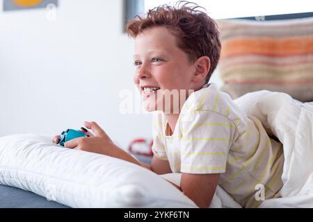 Ein fröhlicher männlicher Spieler sitzt bequem auf einer weißen Couch, während er ein tragbares Videospiel spielt, breit lächelt und von der Kamera wegblickt. Stockfoto