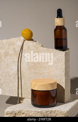 Eine minimalistische Einrichtung mit einer Tropfflasche aus Glas und einem Cremebecher, der stilvoll auf rauen Steinpodesten mit einem getrockneten botanischen Akzent angeordnet ist. Stockfoto
