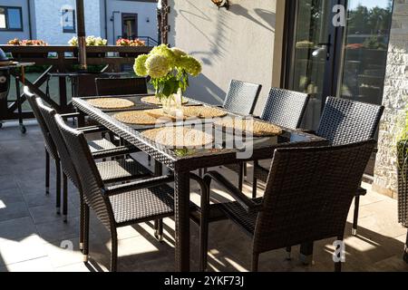 Ein einladender Essbereich im Freien auf einer sonnigen Terrasse mit modernem Korbtisch mit Tischsets und einem frischen Blumenstrauß. Stockfoto