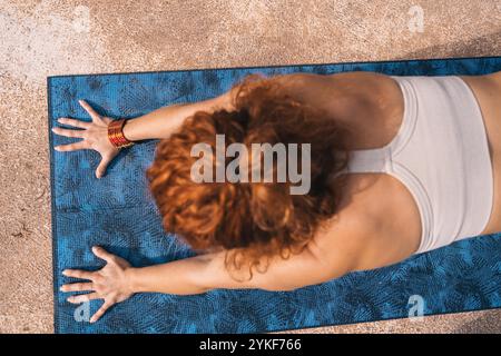 Draufsicht einer zugeschnittenen Rückansicht einer nicht erkennbaren Frau, die eine Yoga-Pose auf einer blauen Matte ausführt. Ihre fokussierte Haltung und das lebendige Zubehör ergänzen Stockfoto