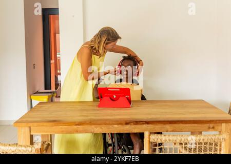 Eine kaukasische Frau passt die Kopfhörer an einen schwarzen Jungen mit Zerebralparese an, der in einem gut beleuchteten Ferienhaus in einem Rollstuhl vor einem Tablet sitzt. Die Stockfoto