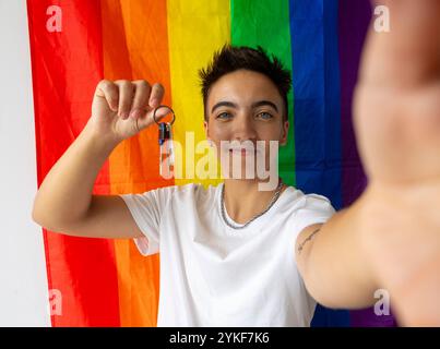 Ein fröhlicher Trans-Mann zeigt seine neuen Hausschlüssel mit einem großen Lächeln, der vor einer lebhaften Stolz-Flagge steht und einen Neuanfang und Stolz auf seine symbolisiert Stockfoto