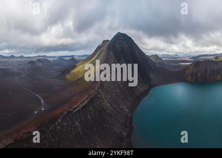 Dramatische Aussicht auf ein Auto, das neben einem tiefblauen vulkanischen See fährt, flankiert von zerklüfteten Bergen im isländischen Hochland unter einem bewölkten Himmel. Stockfoto