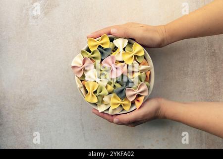 Beschnittene, nicht erkennbare Personenarme mit einer Schüssel farbenfroher Farfalle-Pasta aus Kurkuma, Spinat, Rote Bete, Paprika und Tintenfisch. Der Schuss i Stockfoto