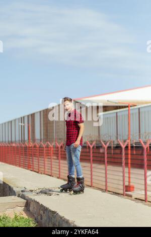 Ein junger Mann in einem roten karierten Hemd und Jeans steht selbstbewusst auf Inline-Skates und blickt von der Kamera weg, vor einem industriellen Hintergrund mit Stockfoto