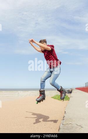 Action-Aufnahme eines jungen Mannes, der auf Inlineskates entlang eines Weges am Meer fährt, ein rotes Flanellhemd und eine Jeans trägt und mit den Armen einen Sprung durchführt Stockfoto