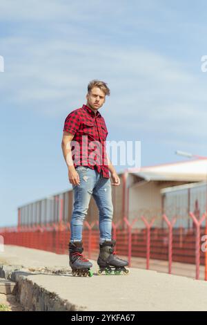 Junger Mann in einem roten karierten Hemd und Denim-Jeans, der auf Inlineskates fährt. Er schaut in die Kamera und steht selbstbewusst in einem urb Stockfoto
