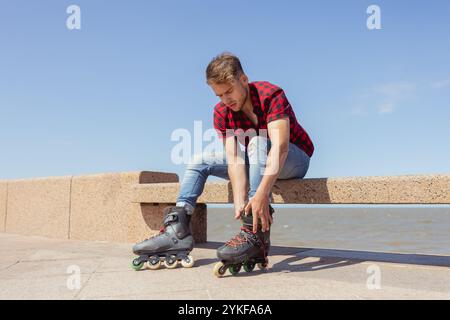 Ein junger Mann wird gezeigt, wie er seine Inlineskates bindet, während er auf einem Steinvorsprung am Meer sitzt und sich auf eine Inlineskates vorbereitet. Er trägt ein R Stockfoto