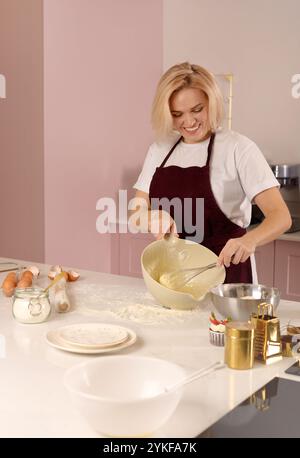 Eine fröhliche Frau mischt in einer stilvollen Küche mit rosa Wänden, umgeben von Backutensilien und Zutaten Stockfoto