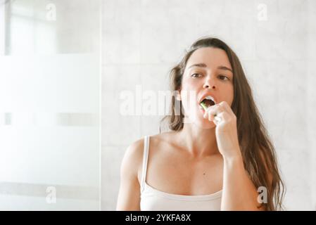 Eine junge Frau in einem weißen Tanktop putzt sich in einem gut beleuchteten Badezimmer die Zähne mit einem fokussierten Ausdruck. Stockfoto
