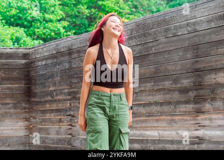 Asiatische Frau mit leuchtend roten Haaren, die fröhlich lächelt, während sie in einer üppigen Umgebung steht, in legeren grünen Hosen und einem schwarzen Oberteil, das die umschließt Stockfoto